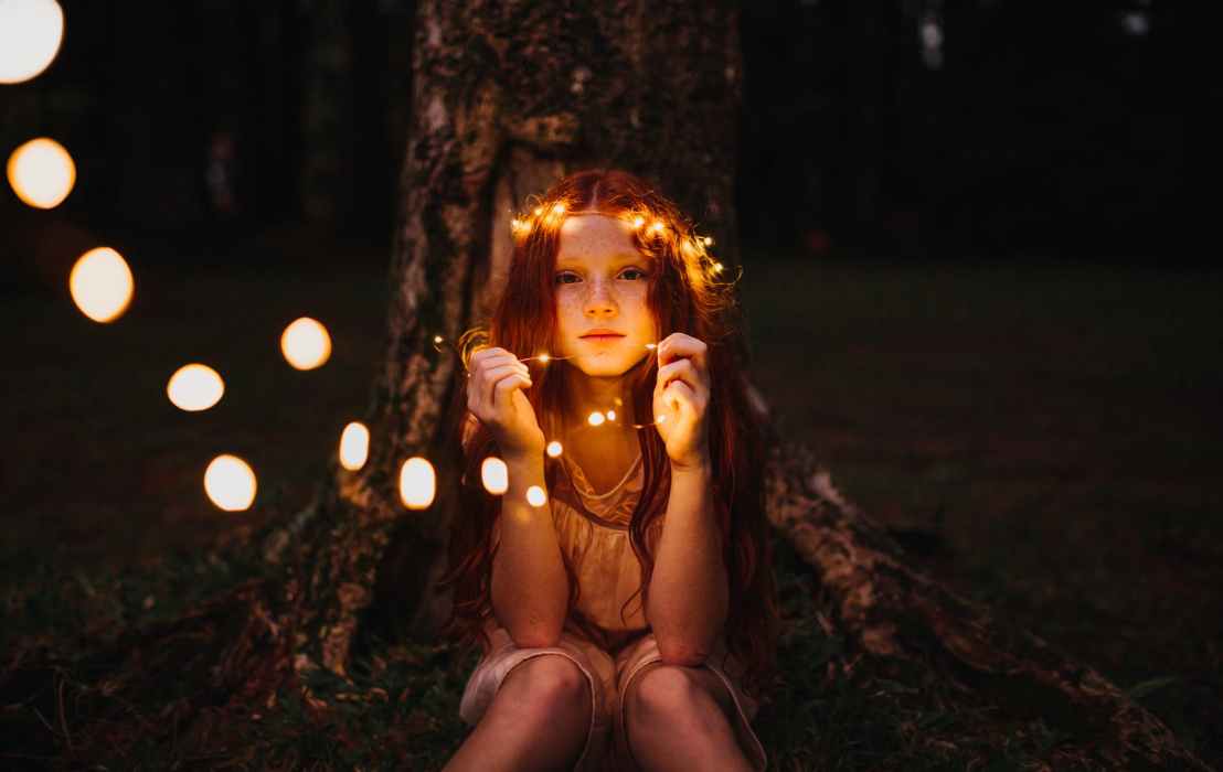 A white kid with long red hair, a string of lights around her head, sitting in front of a tree trunk at night, holding the lights in her hand, looking past the camera. Photo by Matheus Bertelli on Pexels.com