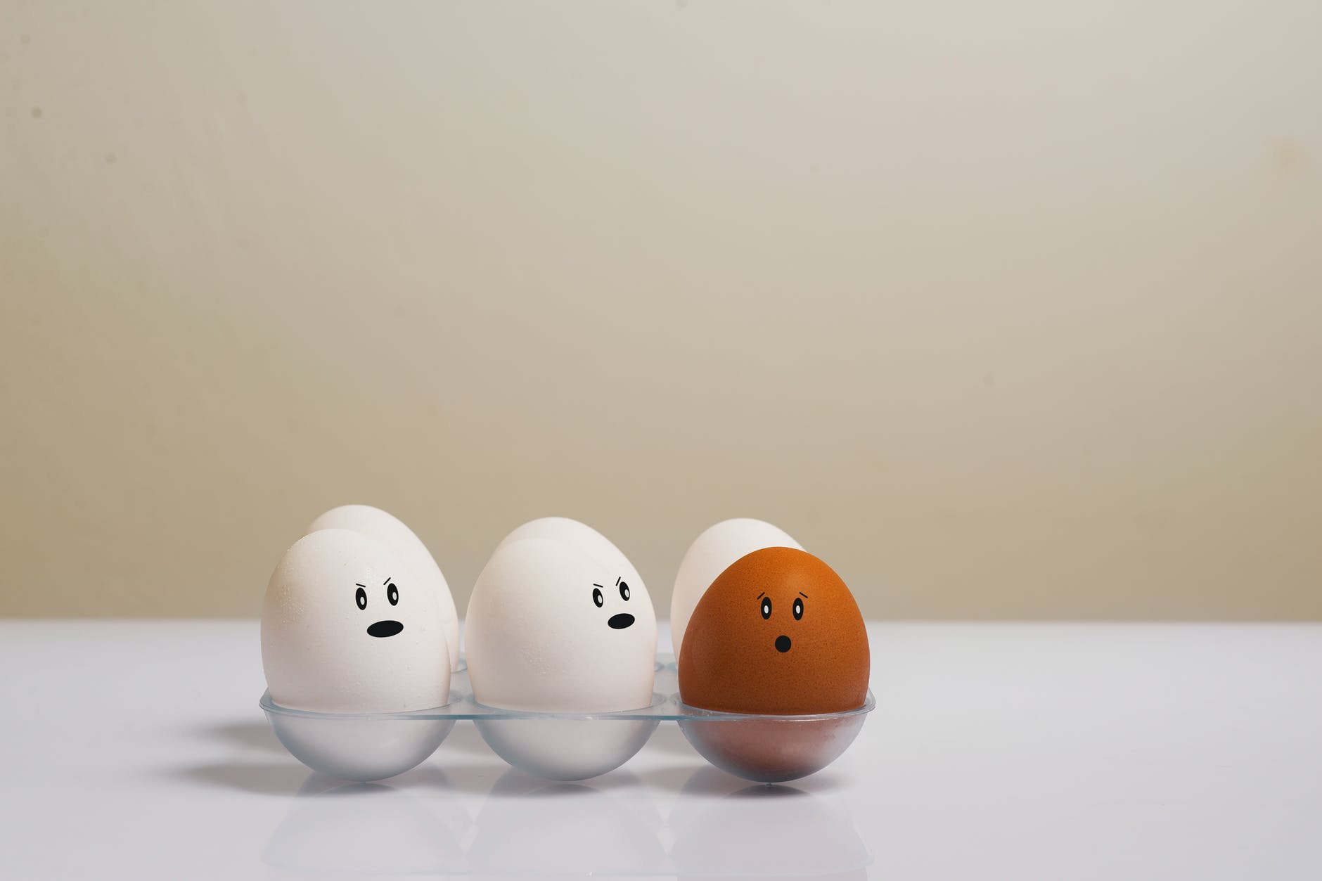 Five white eggs with faces, two looking at one brown egg angrily. The brown egg's mouth is circular and looking at the camera.