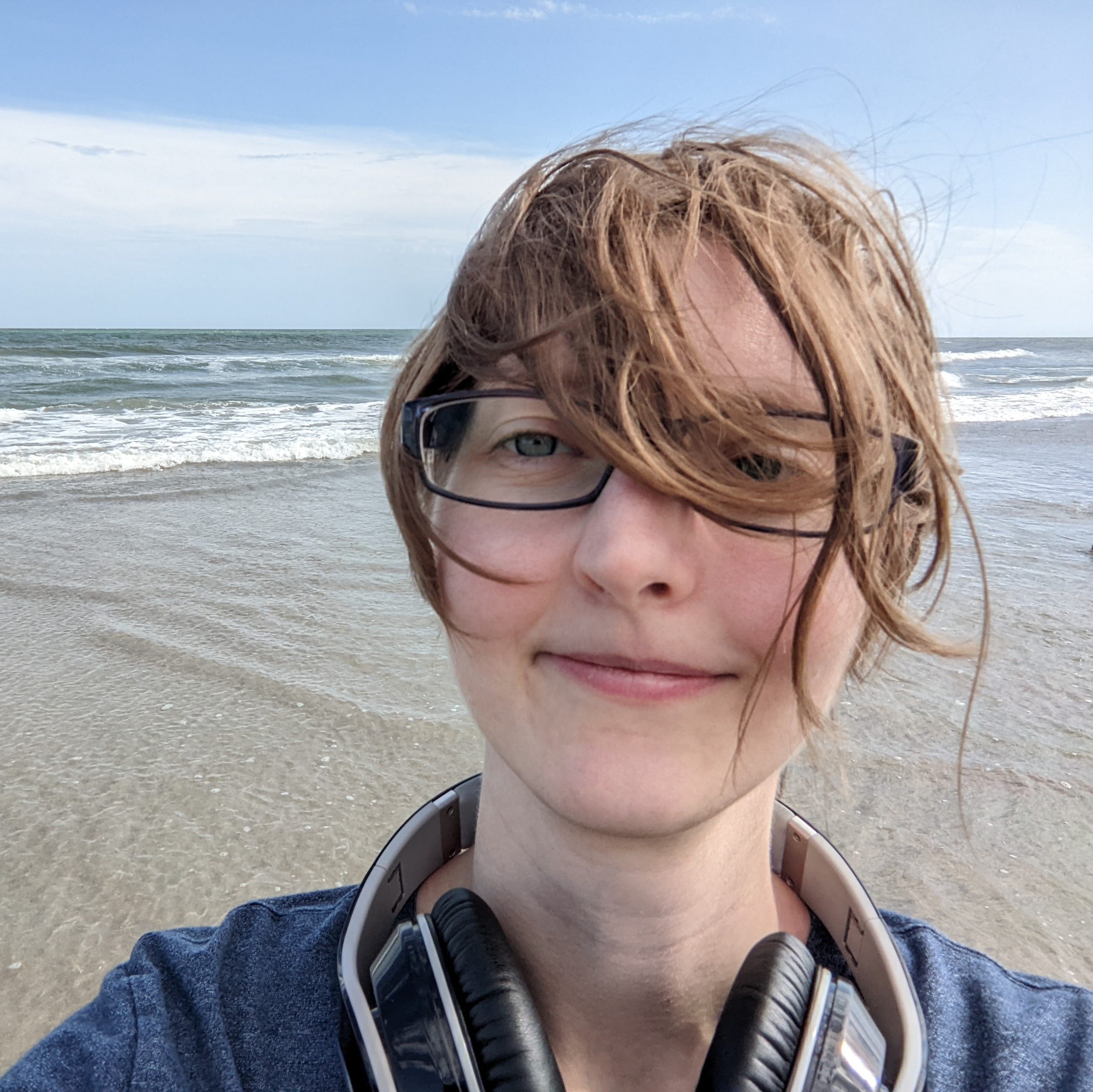 White person at the beach slightly smiling with blond short hair in front of their face, blue eyes, glasses, a navy blue shirt, and grey headphones around their neck. Frothy ocean waves and sand in background.